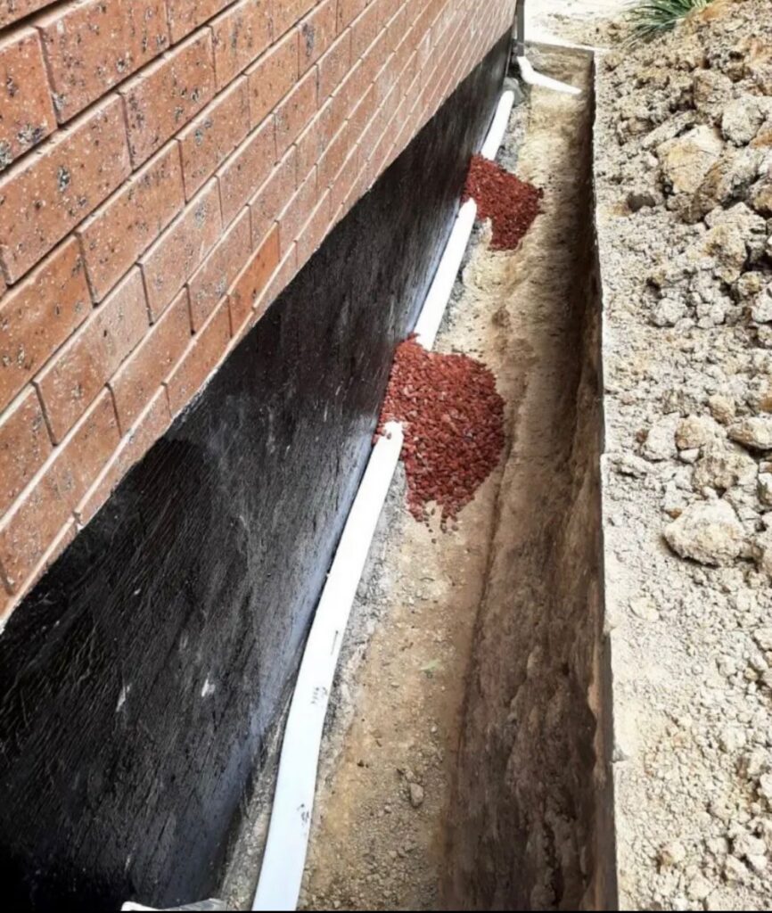 Exterior waterproofing in progress, with a trench excavated alongside a brick foundation to treat the wall from the outside.